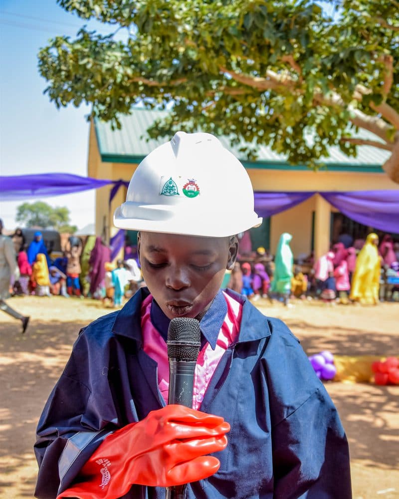 A pupil in engineer's coveralls, hard hat, and work gloves holding a microphone